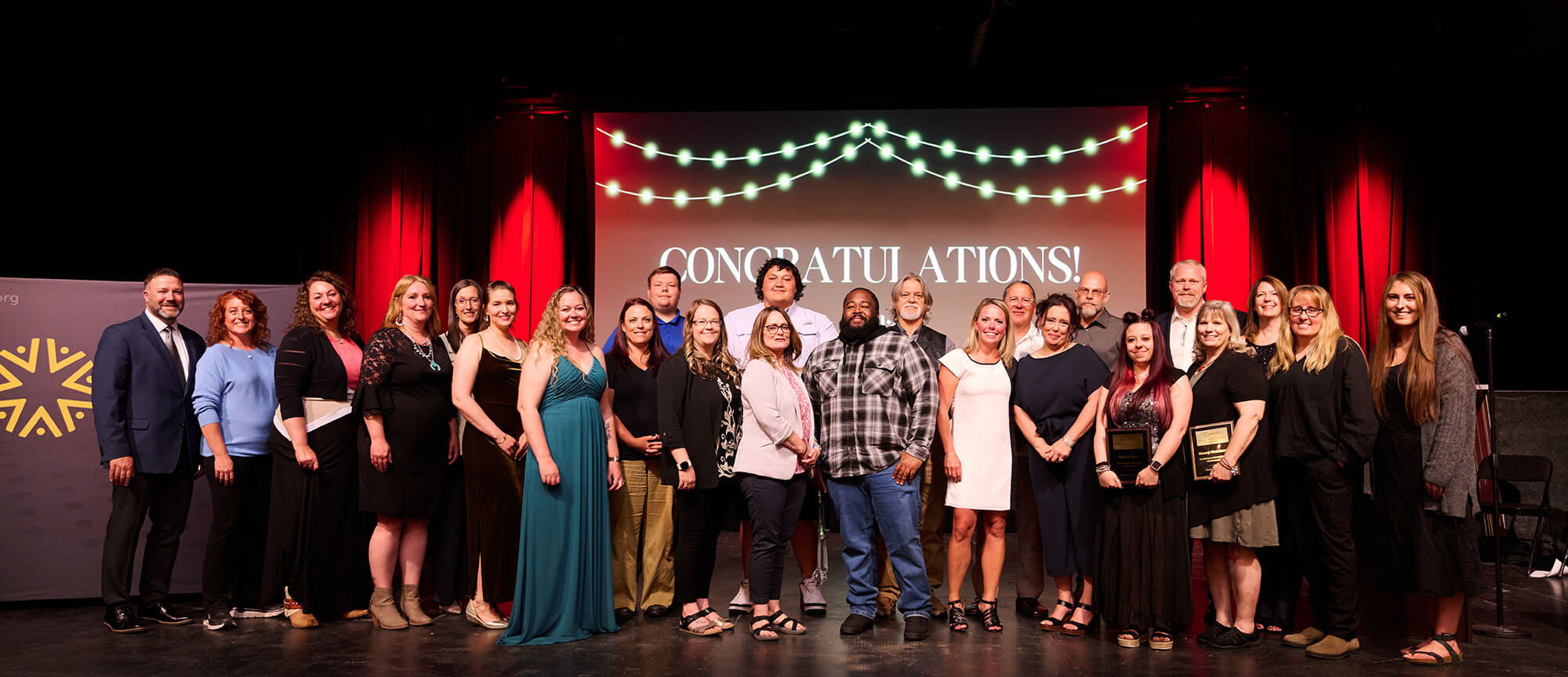 Group of AWARE employees and award winners standing on a stage at the UCC Awards Event 2024. A congratulatory banner reading 'CONGRATULATIONS!' is displayed in the background, illuminated by string lights. The setting features red curtains and spotlights, with attendees dressed in formal and semi-formal attire, smiling proudly for the group photo.