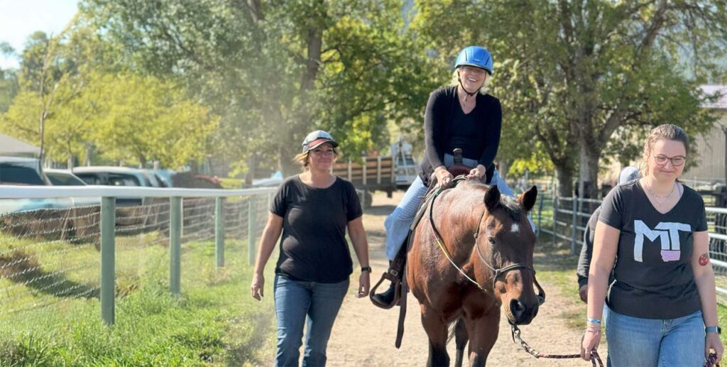 A smiling person wearing a blue helmet is riding a brown horse on a sunny day. They are accompanied by two women walking alongside the horse, one holding its reins. The scene is set on a dirt path surrounded by greenery, with trees and fencing in the background.
